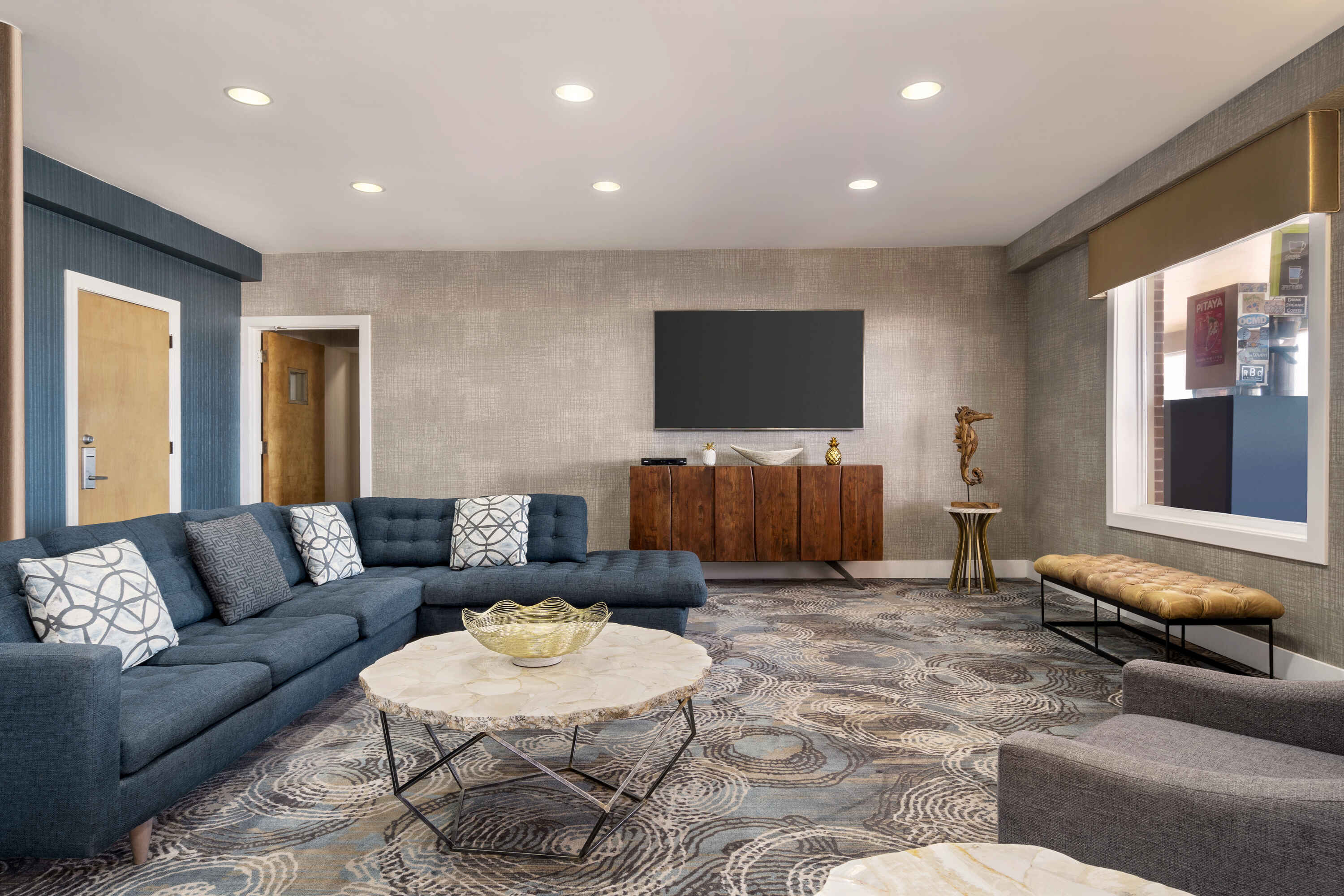 A hotel lobby with a blue sectional sofa, a marble coffee table, a large flat screen TV mounted on the wall, and a wooden credenza underneath it.