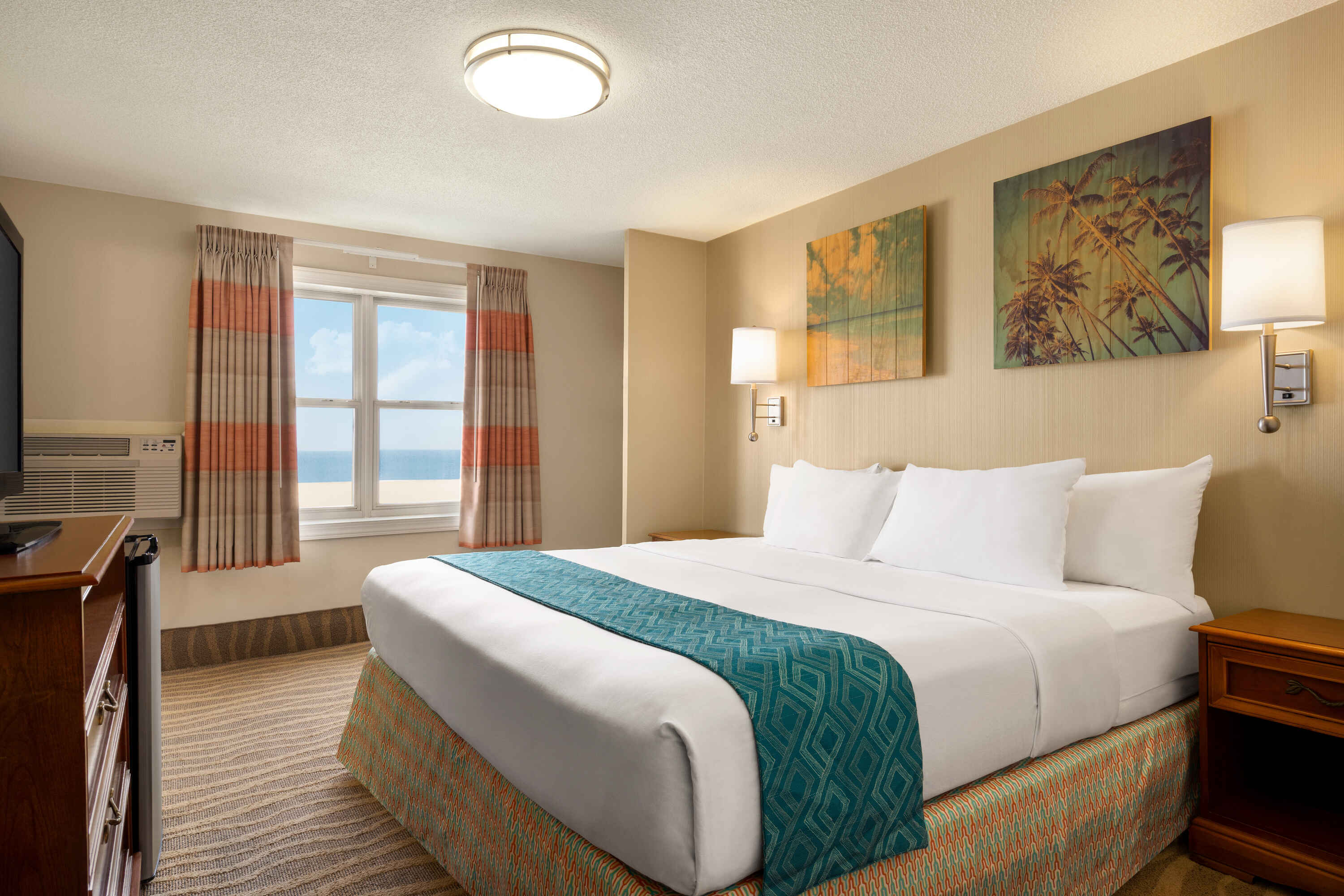 A hotel room with a large bed, window with ocean view, and artwork on the wall.