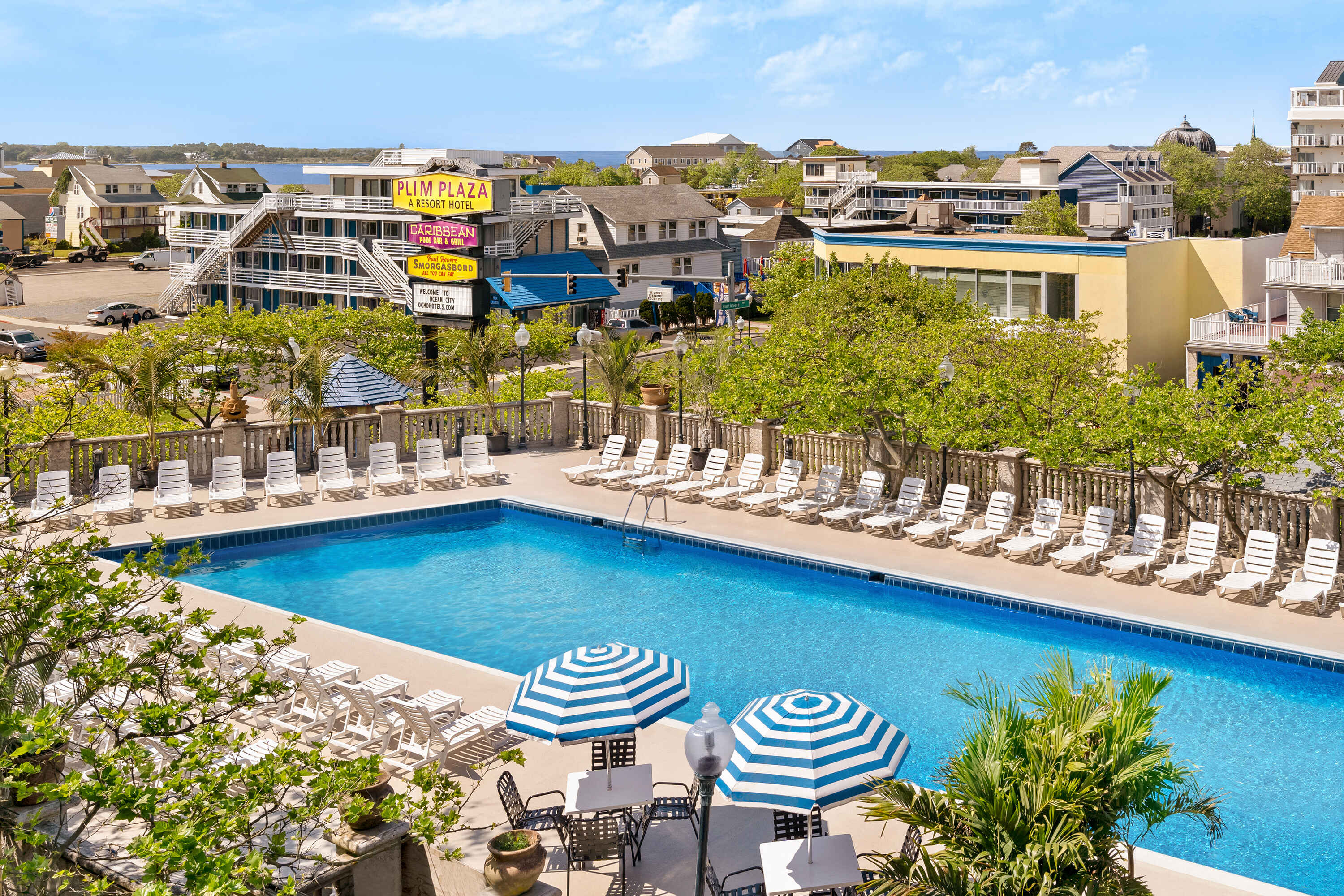 A large pool area with lounge chairs and umbrellas, surrounded by trees and a building in the background.