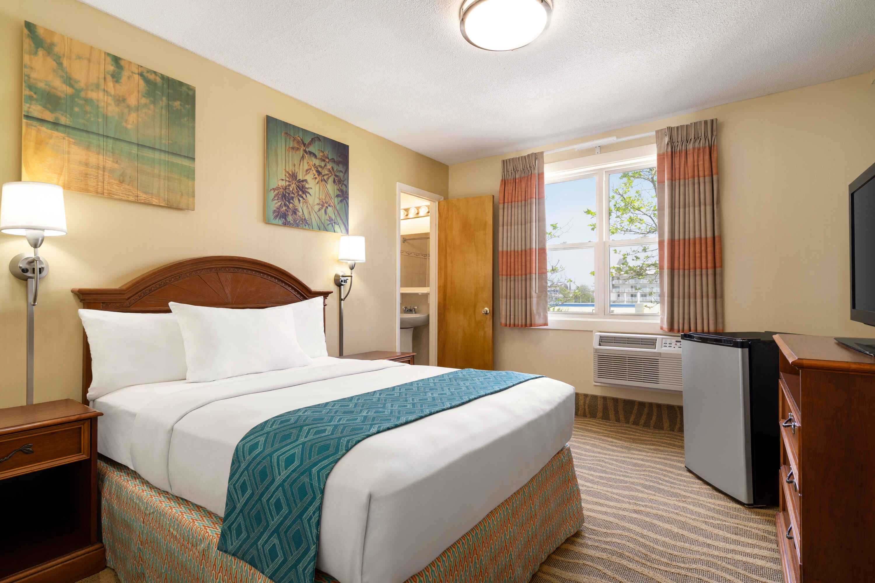 A hotel room with a bed, window, TV, and artwork on the walls.