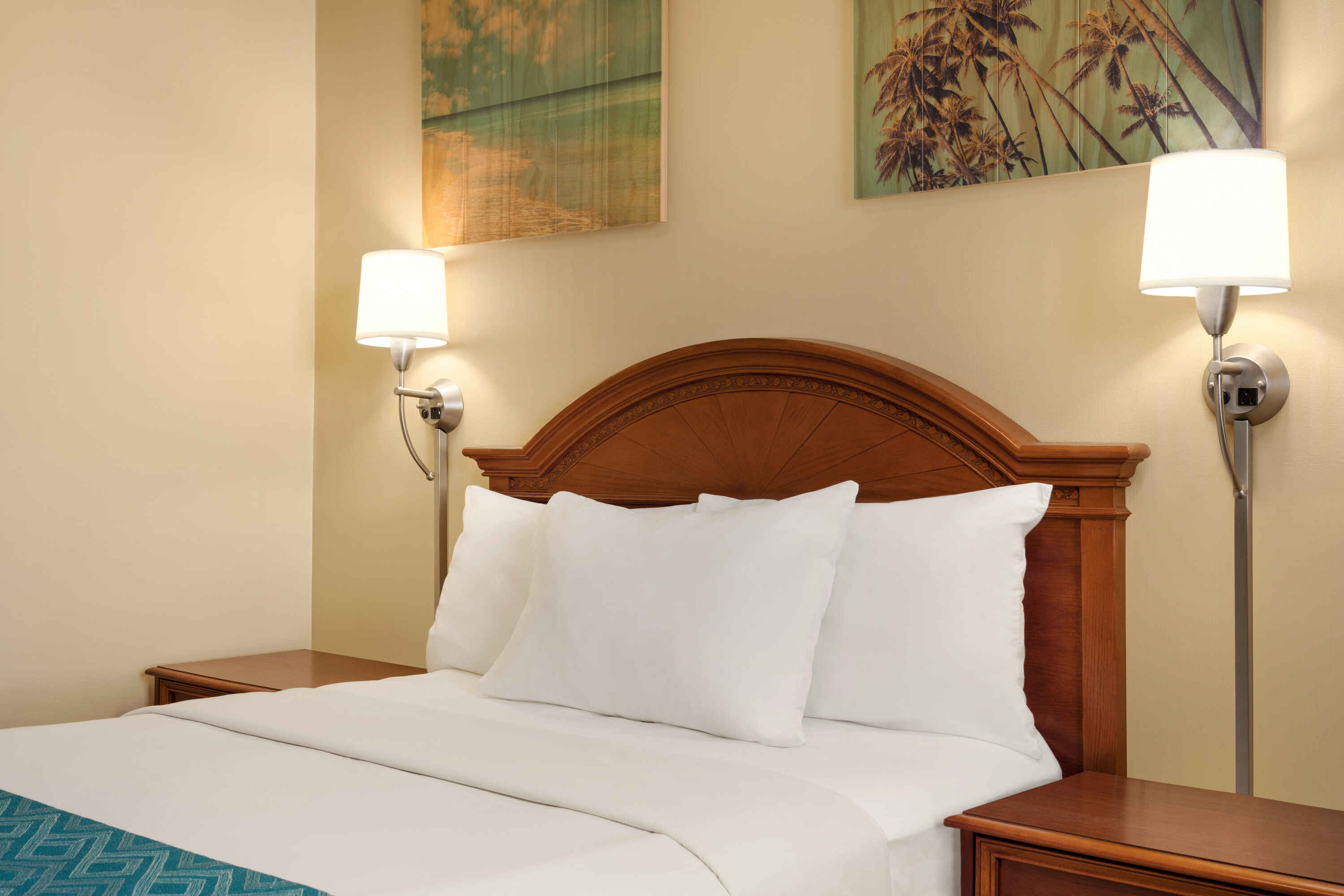 A neatly made bed with a wooden headboard, white bedding, and two nightstands with lamps, against a beige wall with artwork.
