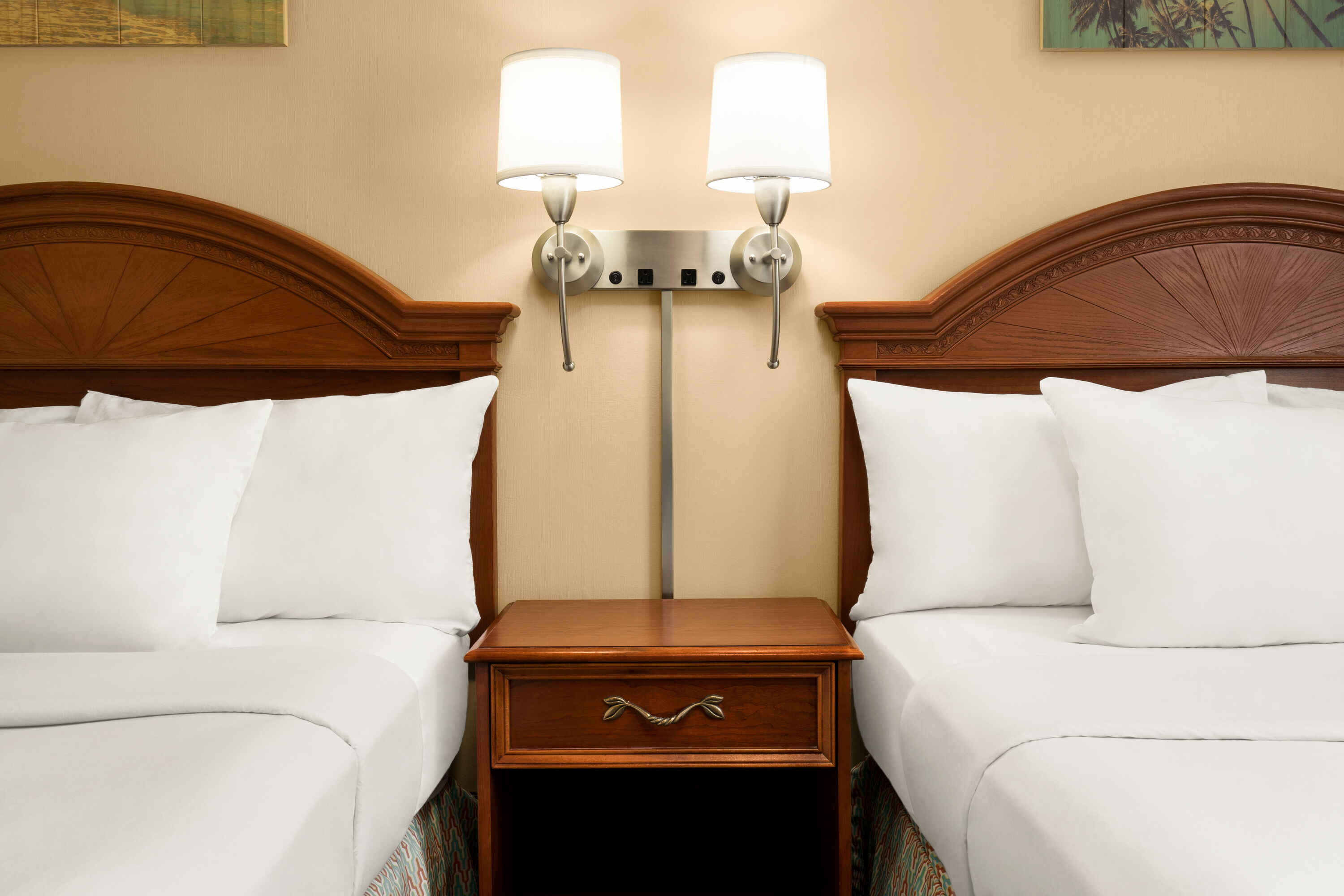 A close-up of two beds with white linens and wooden headboards flanking a nightstand with a lamp.